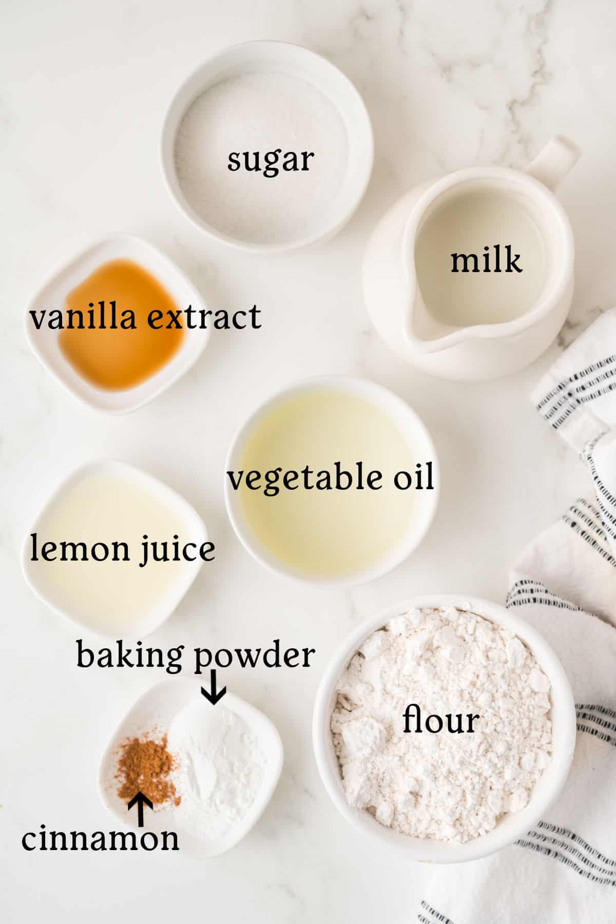 Bowls and a pitcher containing labeled ingredients: sugar, milk, vanilla extract, lemon juice, vegetable oil, flour, baking powder, and cinnamon on a white surface.