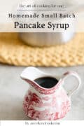 Small red and white pitcher filled with pancake syrup on a countertop, with a saucepan and woven mat in the blurred background. Text overlay reads Homemade Small Batch Pancake Syrup.