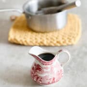 A small patterned ceramic pitcher filled with homemade pancake syrup sits on a counter, with a saucepan and whisk on a yellow trivet blurred in the background.