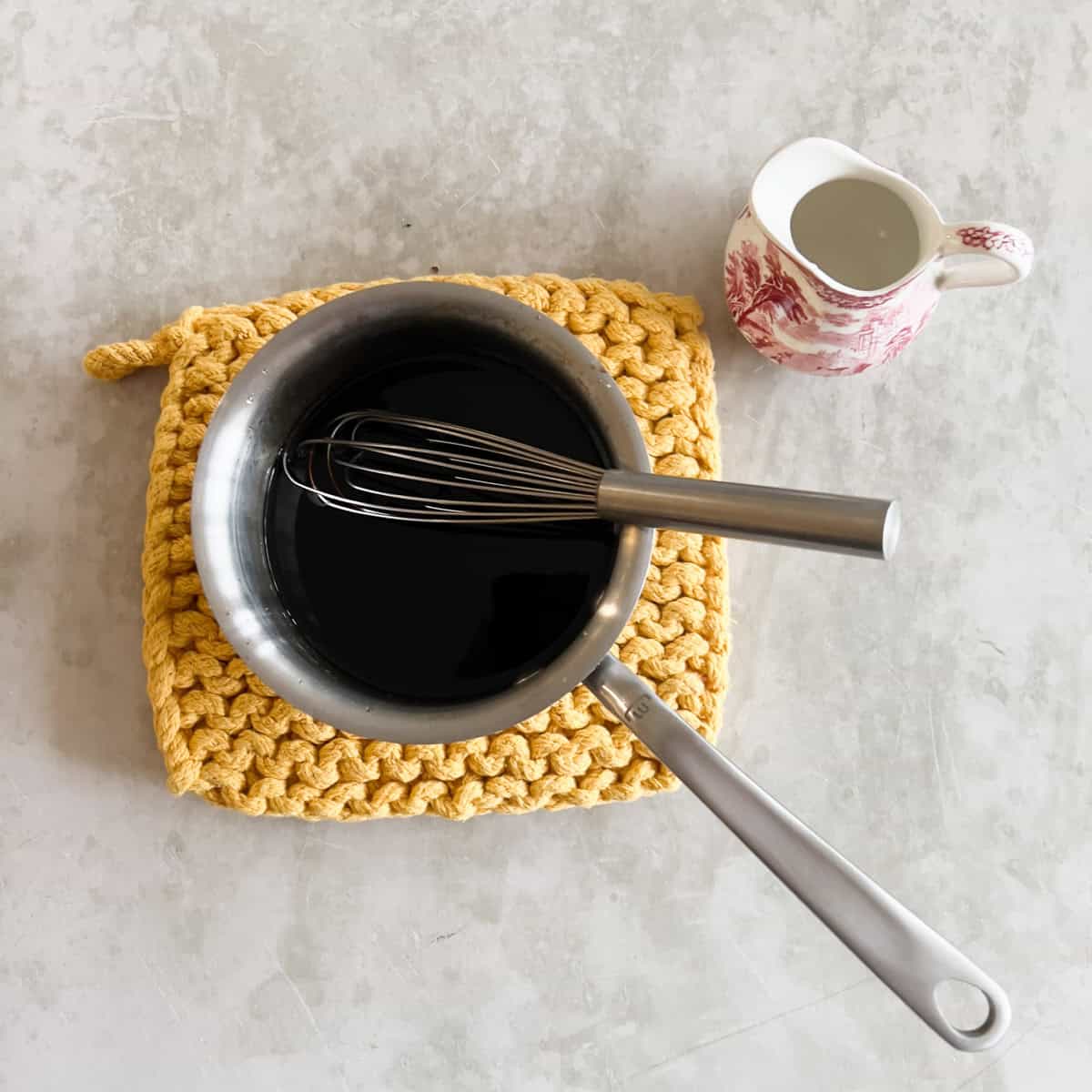 A saucepan with a whisk inside, filled with homemade pancake syrup sits on a yellow knitted trivet next to a small pitcher of water on a light-colored surface.