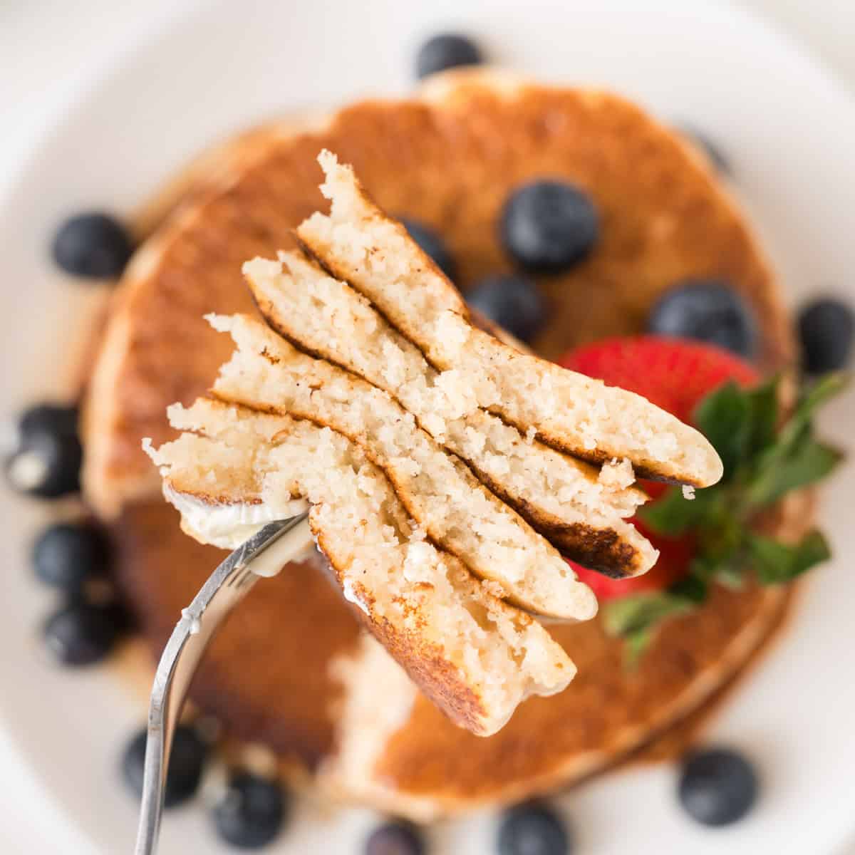 A fork holds a cut section of pancakes above a plate topped with blueberries and a strawberry.