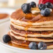 A stack of pancakes topped with blueberries, a pat of butter, and syrup on a white plate.