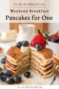 A plate of four pancakes with a bite taken out, topped with whipped cream, blueberries, and a strawberry, with more berries and butter in the background. Text reads “Pancakes for One.”.