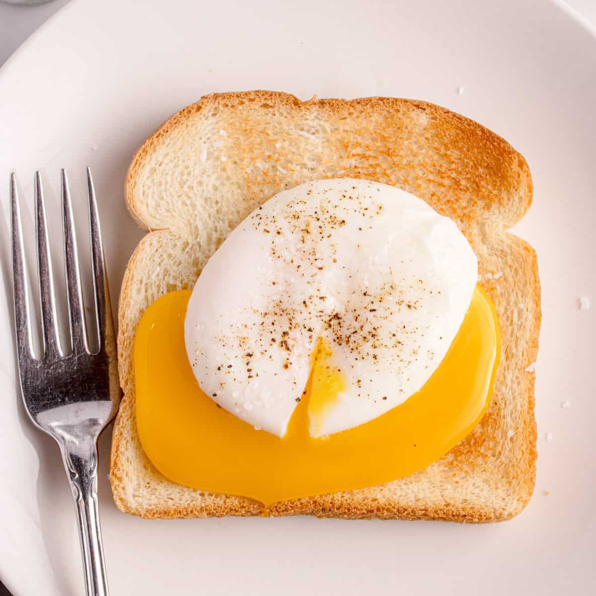 A slice of toasted bread topped with a poached egg, cut open to reveal a runny yolk, on a white plate with a fork.