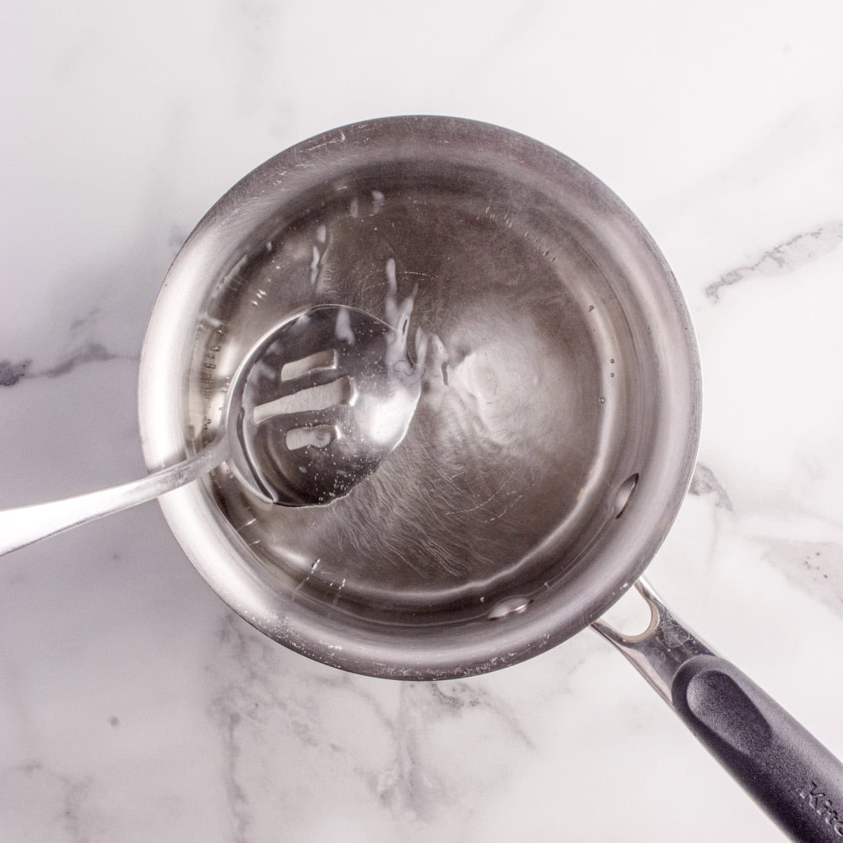 A metal saucepan containing clear liquid with a slotted spoon resting inside, placed on a white marble surface.