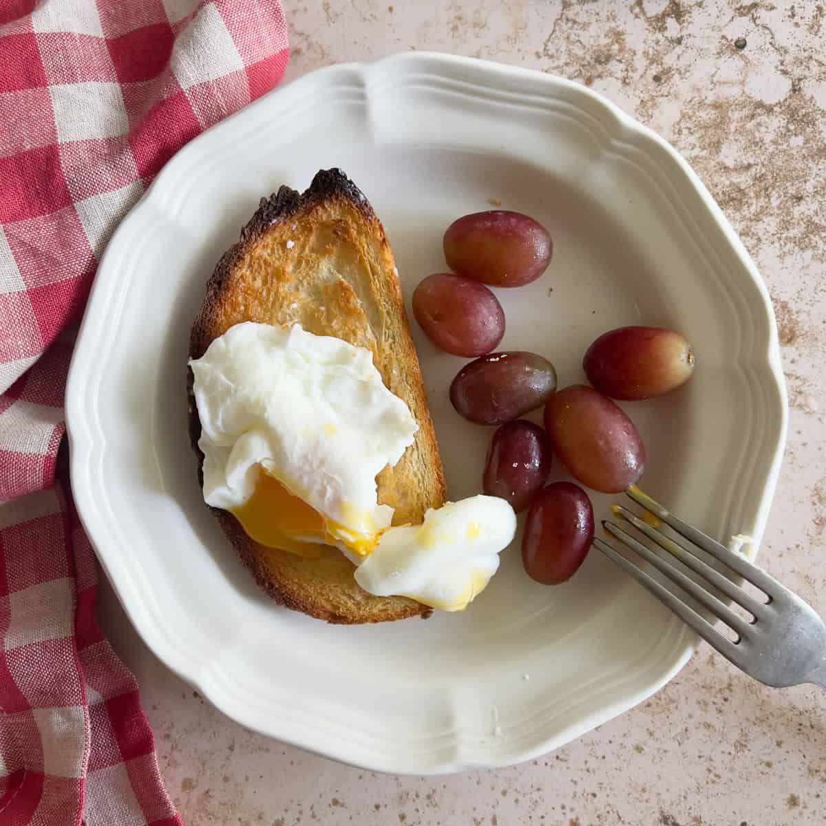 A plate with a slice of toast topped with a poached egg and a side of red grapes. A fork and a red checkered napkin are nearby.