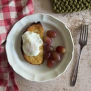A plate with a slice of toasted bread topped with a poached egg, a handful of red grapes, a fork, and a red checkered napkin on the side.