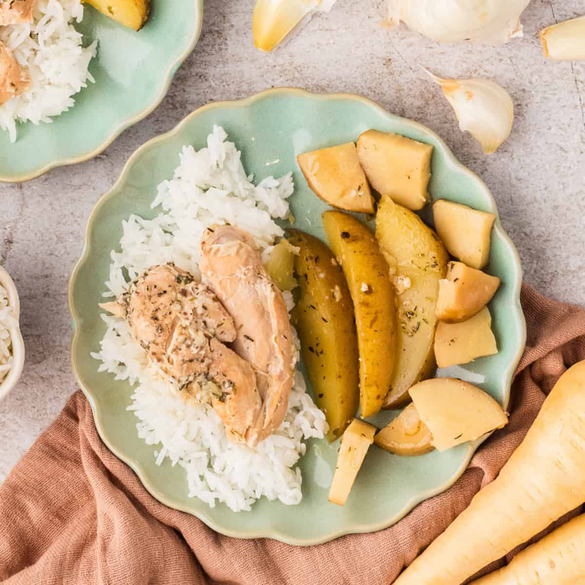 A plate with white rice, a piece of cooked garlic chicken, and roasted potato wedges. Garlic cloves and a parsnip are nearby on the table.