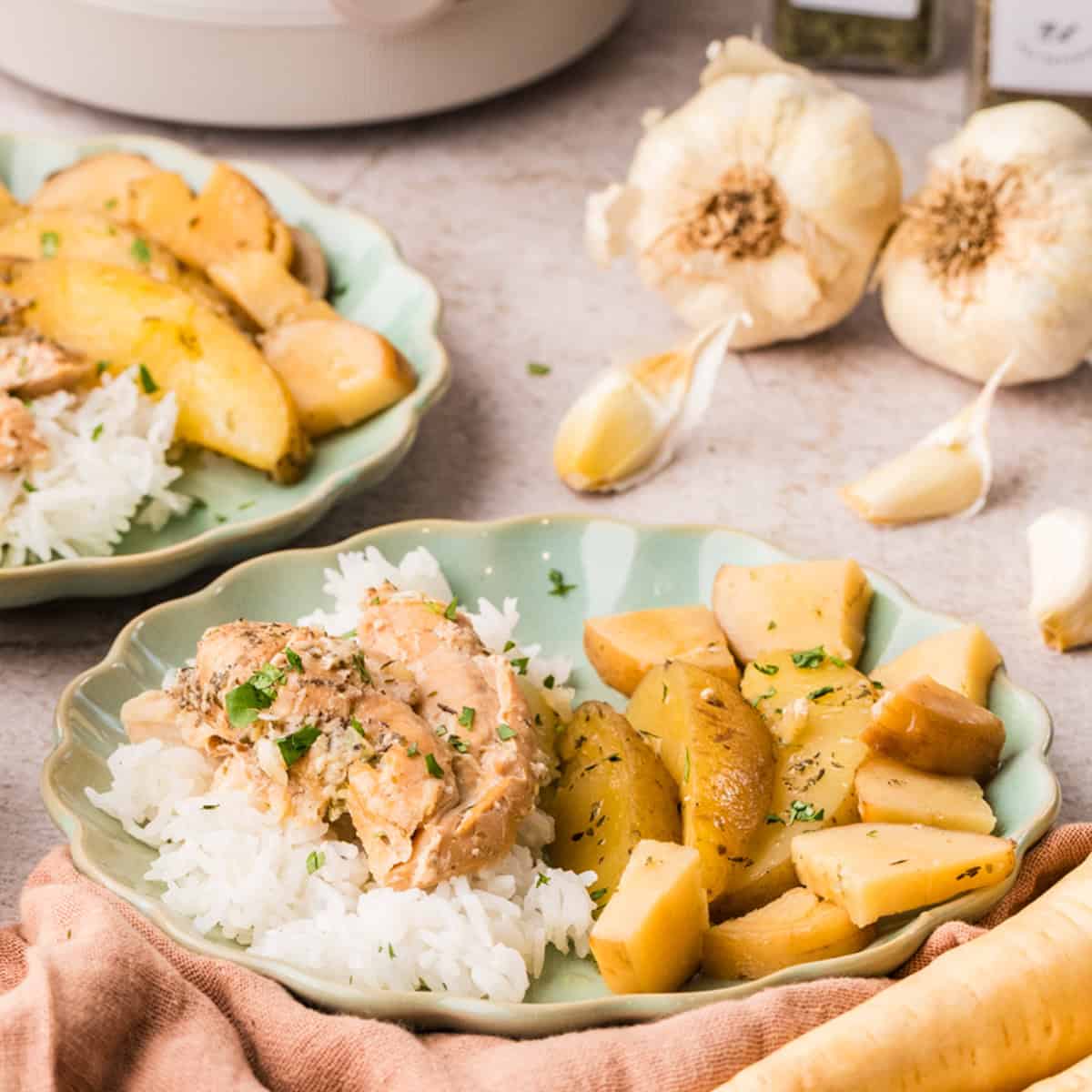 A plate of white rice topped with seasoned chicken and roasted potato wedges, with more potatoes and garlic cloves in the background.