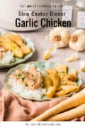 Two plates of garlic chicken with potatoes and white rice are served on a table. Whole garlic bulbs and spices are in the background. Text reads Slow Cooker Dinner: Garlic Chicken.