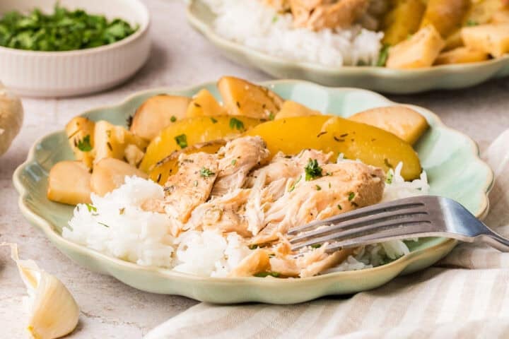 A plate of white rice topped with shredded chicken, cooked potatoes, and herbs, with a fork resting on the food. A bowl of chopped herbs and garlic are in the background.