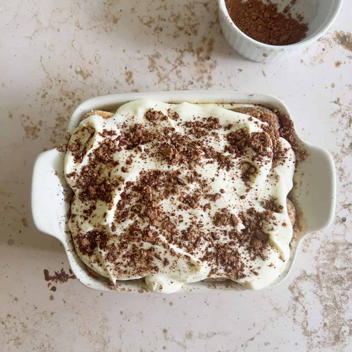 A rectangular dish of tiramisu topped with mascarpone cream and cocoa powder sits on a light marble surface next to a small bowl of cocoa powder.