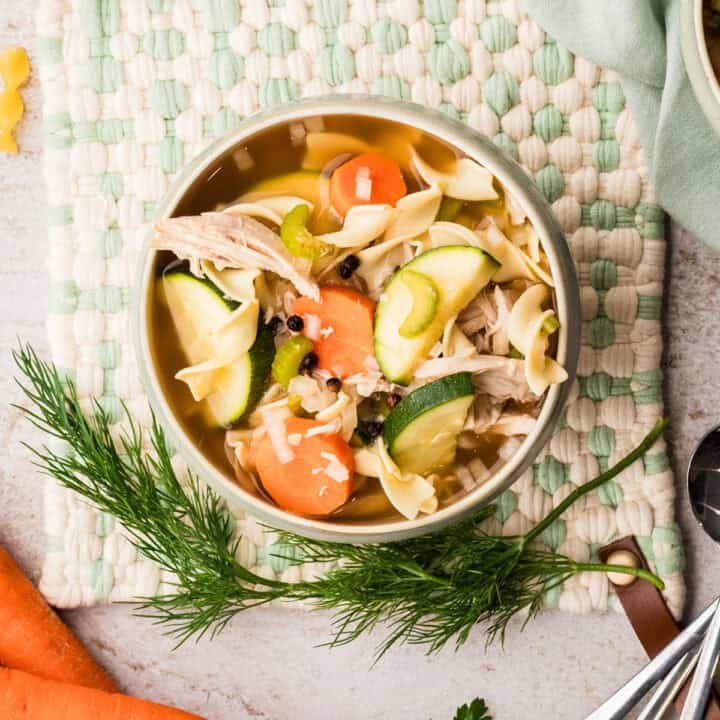 A bowl of chicken noodle soup with carrots, zucchini, celery, and herbs, placed on a woven mat with fresh dill and carrots nearby.