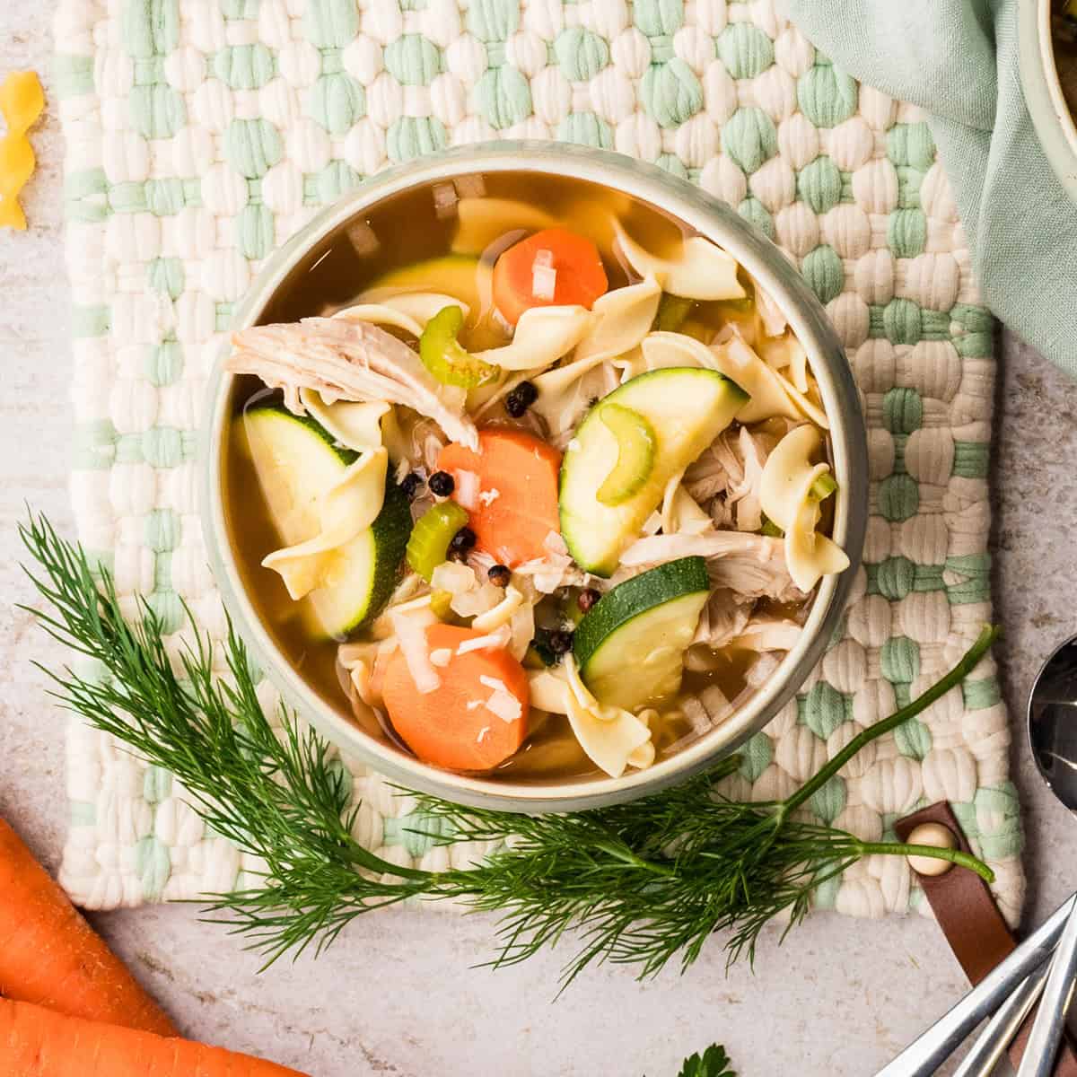 A bowl of chicken noodle soup with carrots, zucchini, celery, and herbs, placed on a woven mat with fresh dill and carrots nearby.
