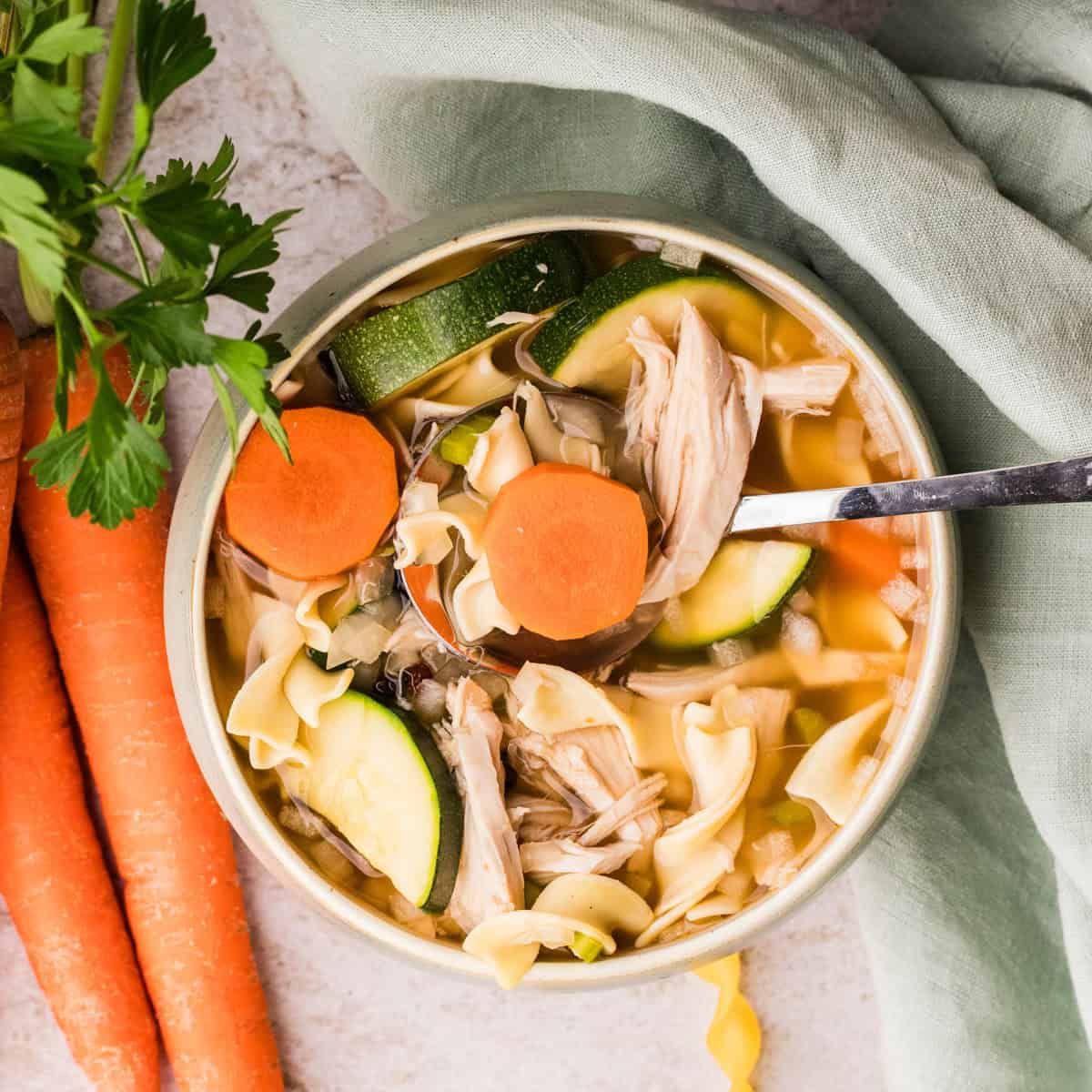 A bowl of chicken noodle soup with sliced carrots and zucchini, next to whole carrots and a sprig of parsley on a light surface.