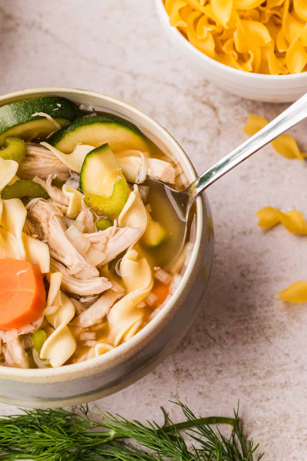 A bowl of chicken noodle soup with carrots, zucchini, and egg noodles, with a spoon inside. A bowl of dry egg noodles and fresh dill are nearby.