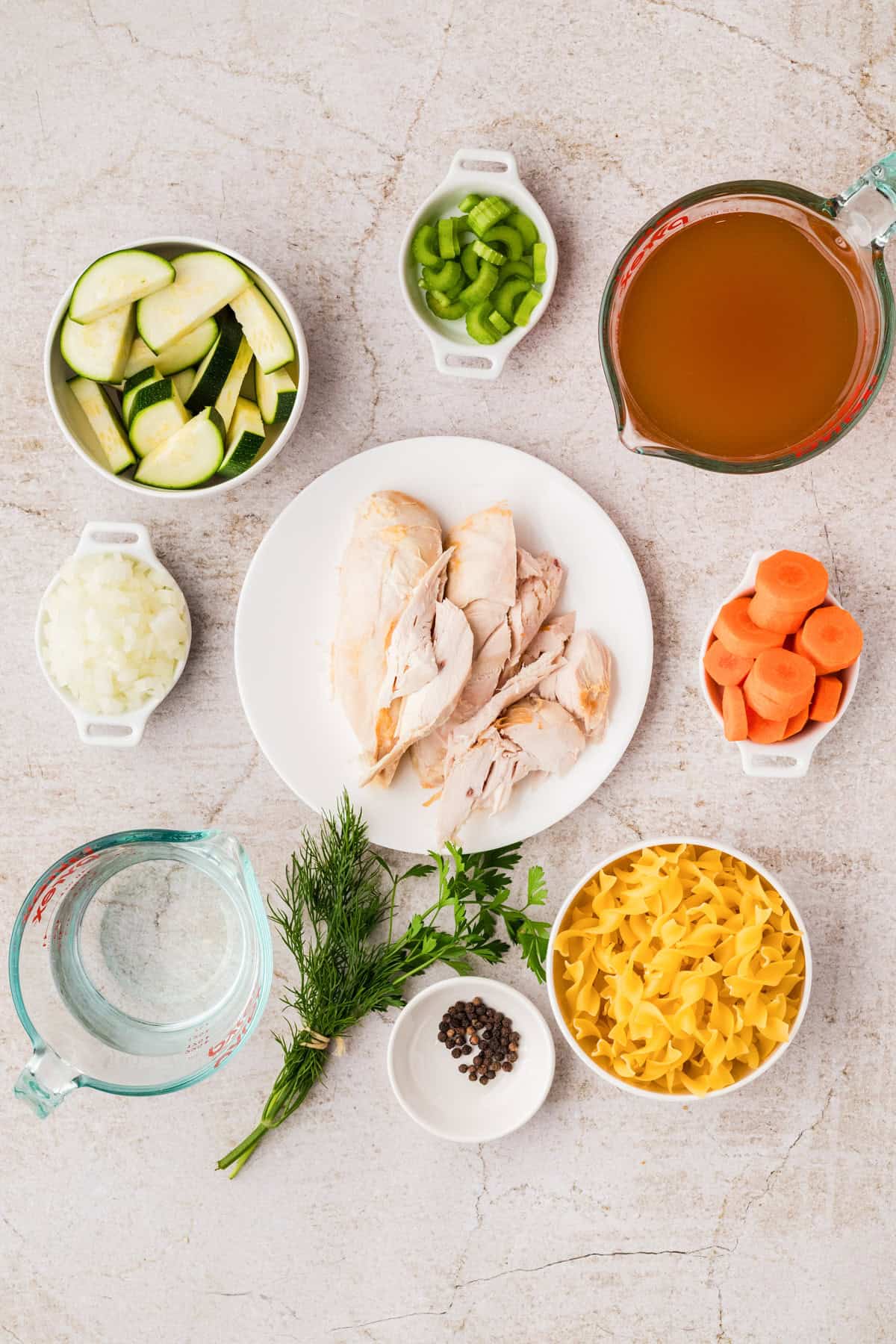 Top-down view of ingredients for chicken noodle soup, including cooked chicken, noodles, carrots, zucchini, onion, celery, broth, water, peppercorns, and fresh herbs on a light surface.