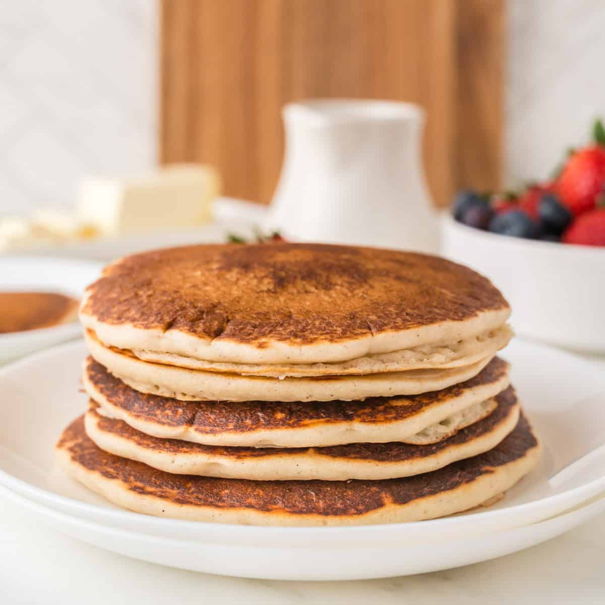 A stack of four pancakes on a white plate with butter, syrup, and a bowl of mixed berries in the background.