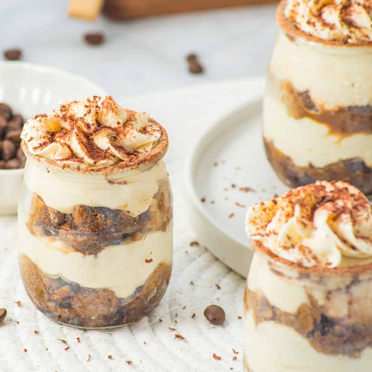 Three jars filled with layers of creamy tiramisu, cake pieces, and topped with whipped cream and cocoa powder, placed on a white surface with scattered coffee beans.