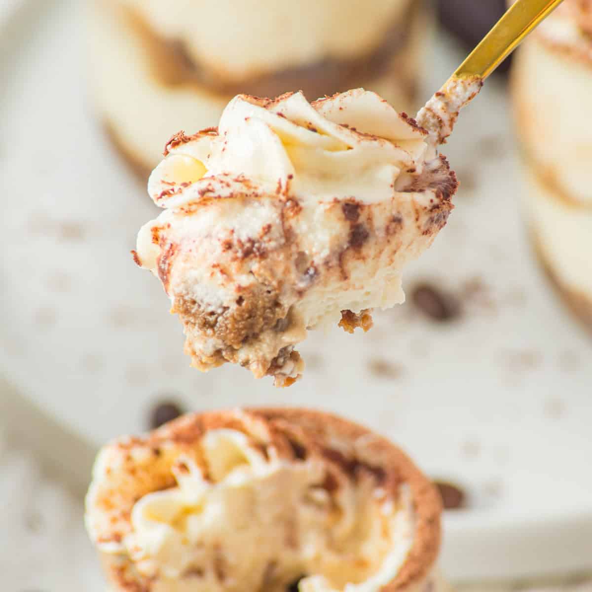 A close-up of a spoonful of creamy tiramisu with layers of mascarpone and cocoa powder, held above a plate with more tiramisu in the background.