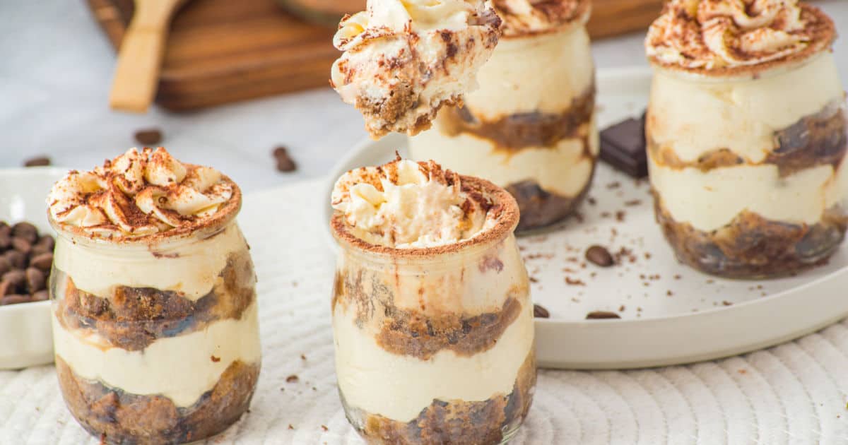 Four jars filled with layered tiramisu, topped with whipped cream and cocoa powder, are arranged on a white tray. A spoon holds a serving above one jar. Coffee beans are scattered nearby.