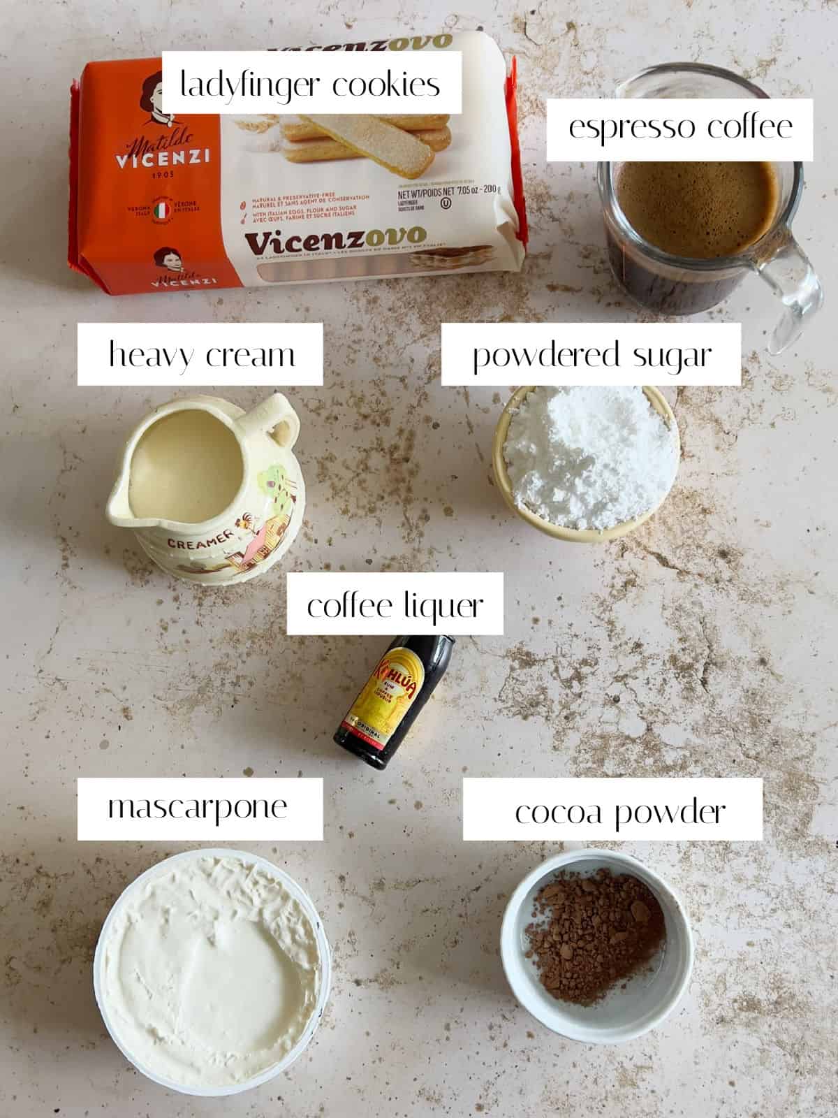 Ingredients for tiramisu are laid out and labeled: ladyfinger cookies, espresso coffee, heavy cream, powdered sugar, coffee liqueur, mascarpone, and cocoa powder.