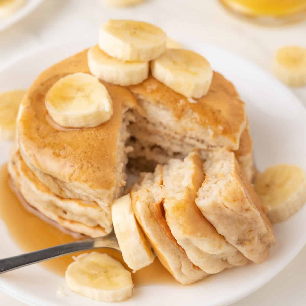 A stack of pancakes topped with banana slices, partially cut with a fork, and surrounded by more banana slices on a white plate.