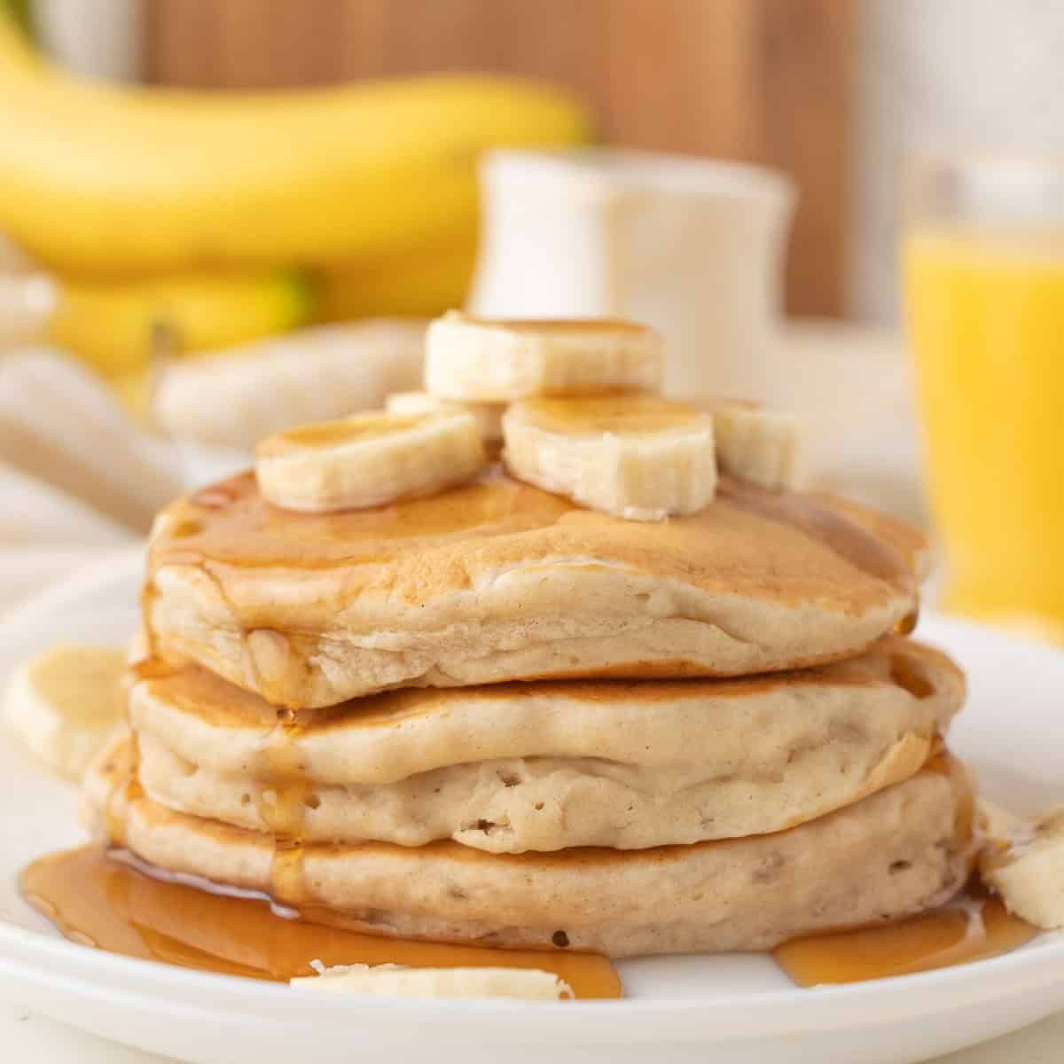 A stack of pancakes topped with banana slices and syrup on a white plate, with bananas and a glass of orange juice in the background.