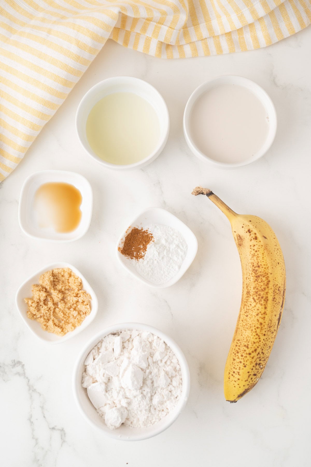 A ripe banana, flour, brown sugar, baking powder, cinnamon, vanilla extract, oil, and milk are arranged in small bowls on a marble surface with a yellow striped cloth.