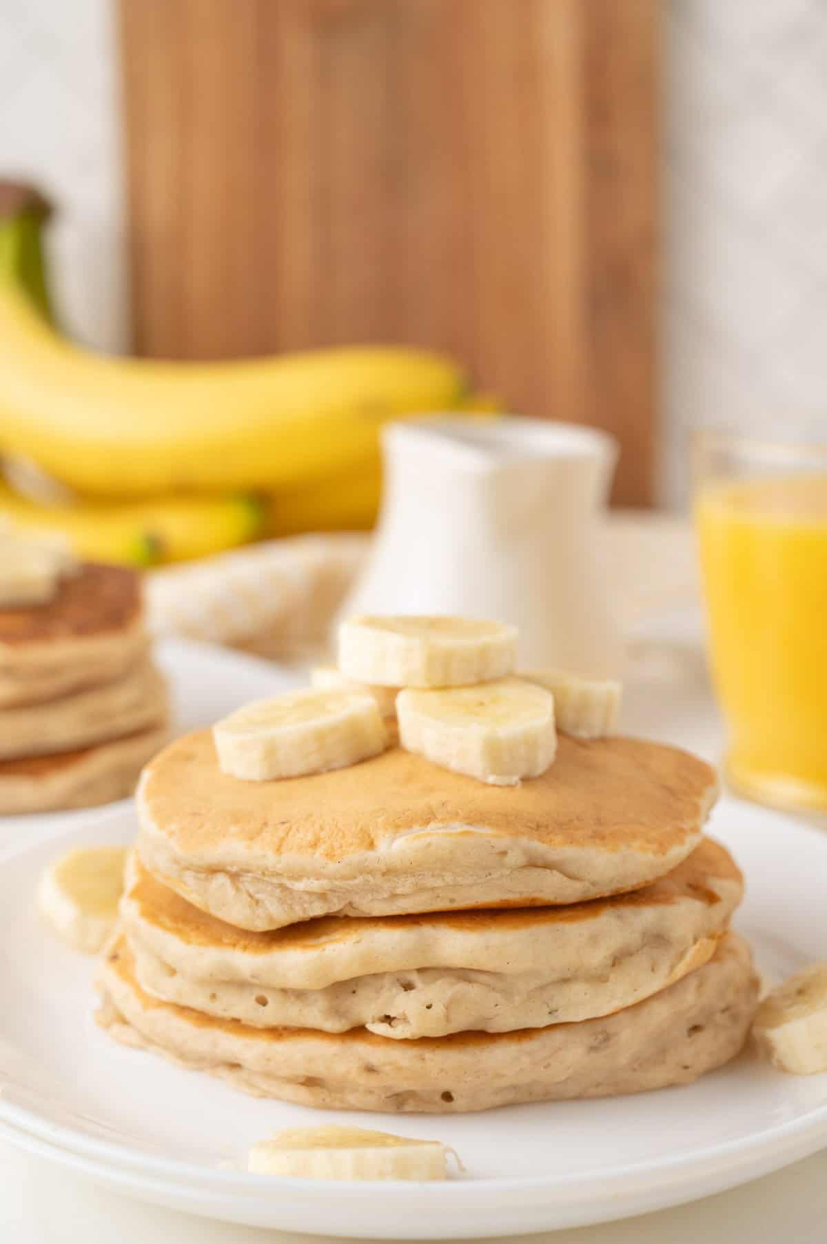 A stack of fluffy banana pancakes topped with banana slices on a white plate, perfect for a single serving, with a glass of orange juice, bananas, and a syrup pitcher in the background.