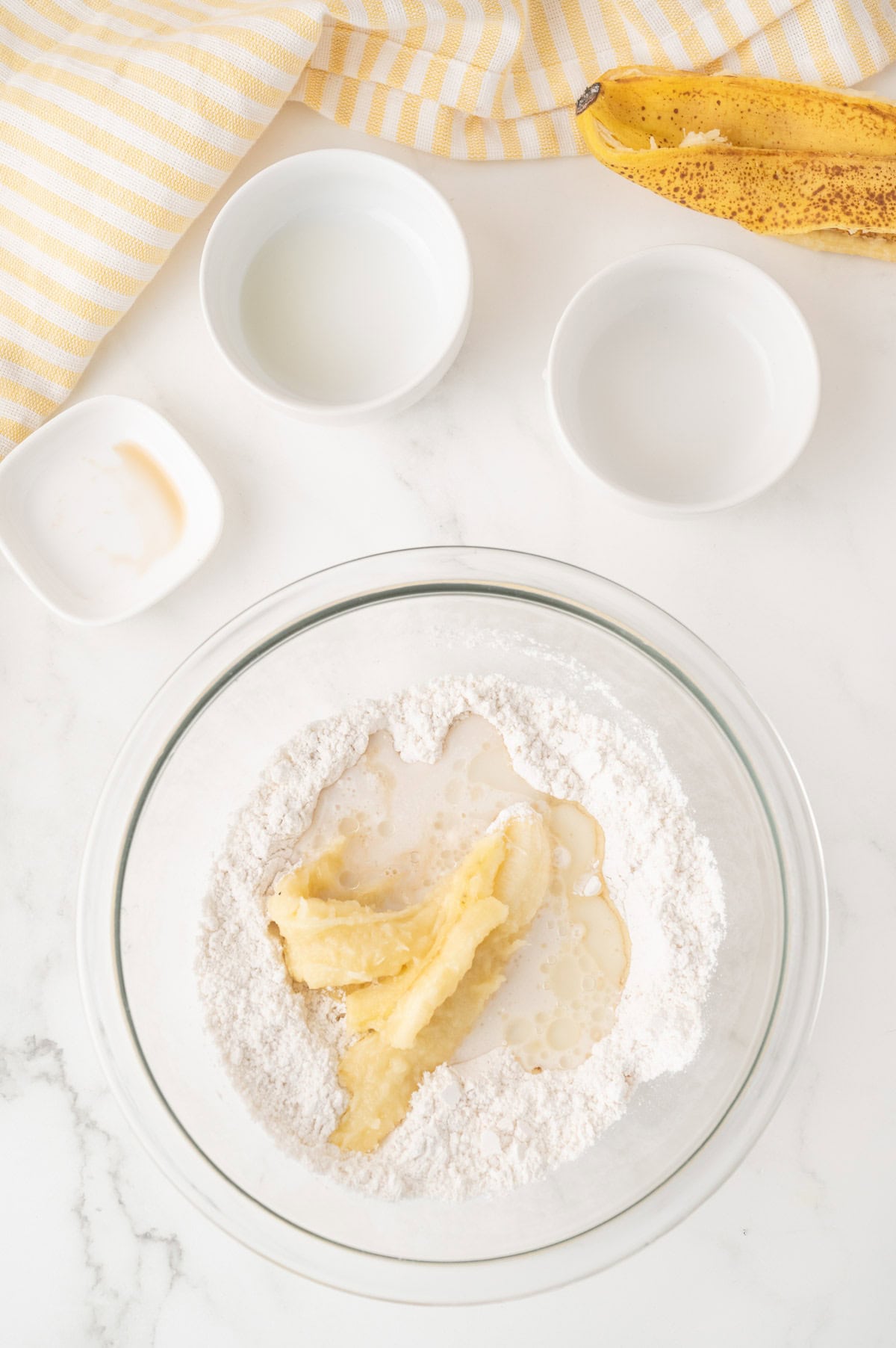 A glass bowl with flour, mashed banana, and liquid ingredients, surrounded by small bowls, a ripe banana, and a yellow-striped towel on a white surface.