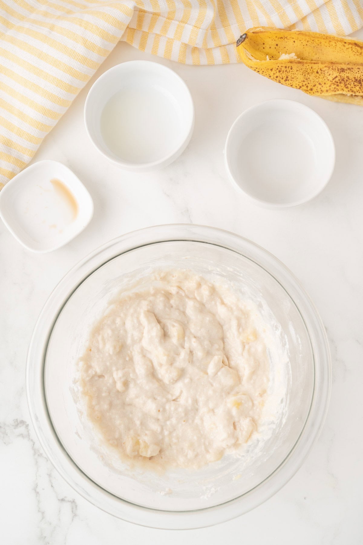 A glass bowl of banana bread batter sits on a white countertop beside two small bowls of liquid, a ripe banana, and a yellow striped towel.
