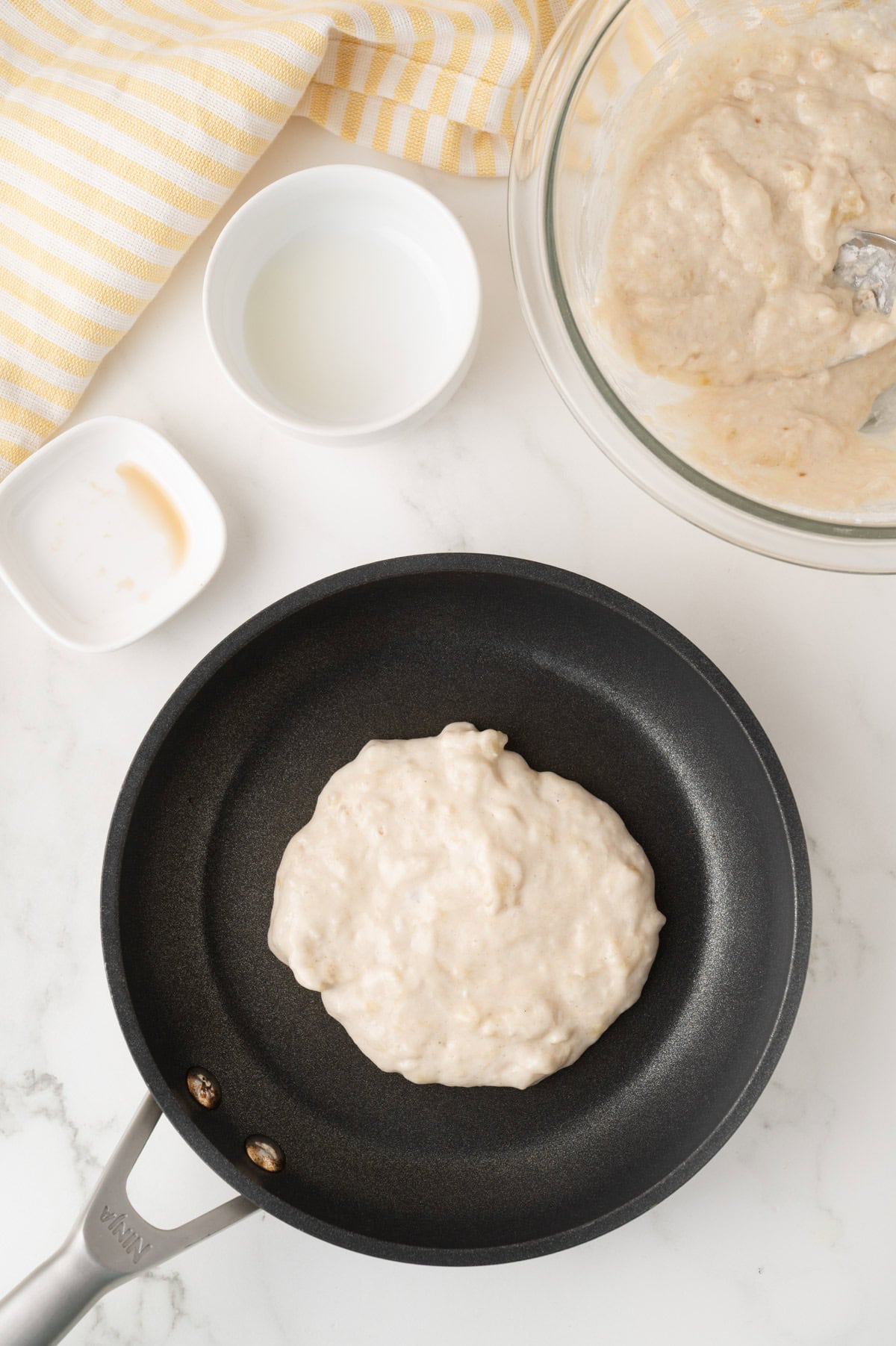 A nonstick pan with uncooked pancake batter, a glass bowl of more batter, a small bowl of oil, and a striped yellow towel on a white surface.