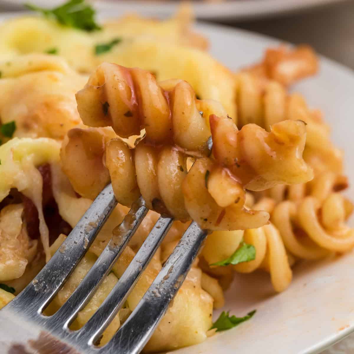 A close-up of a fork holding a bite of rotini pasta with sauce and herbs, with more pasta on a white plate in the background.