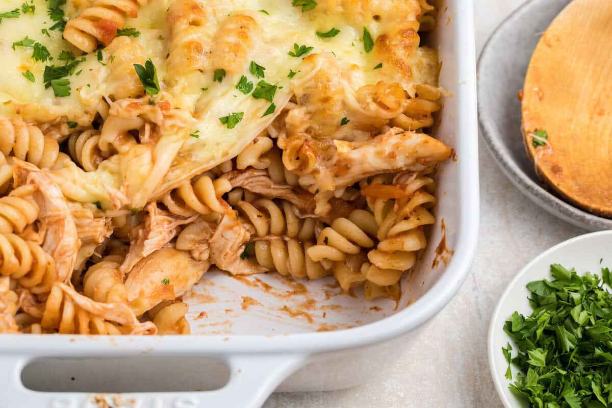 Baked rotini pasta with shredded chicken and melted cheese in a white casserole dish, garnished with chopped parsley. A wooden spoon and a bowl of parsley are nearby.