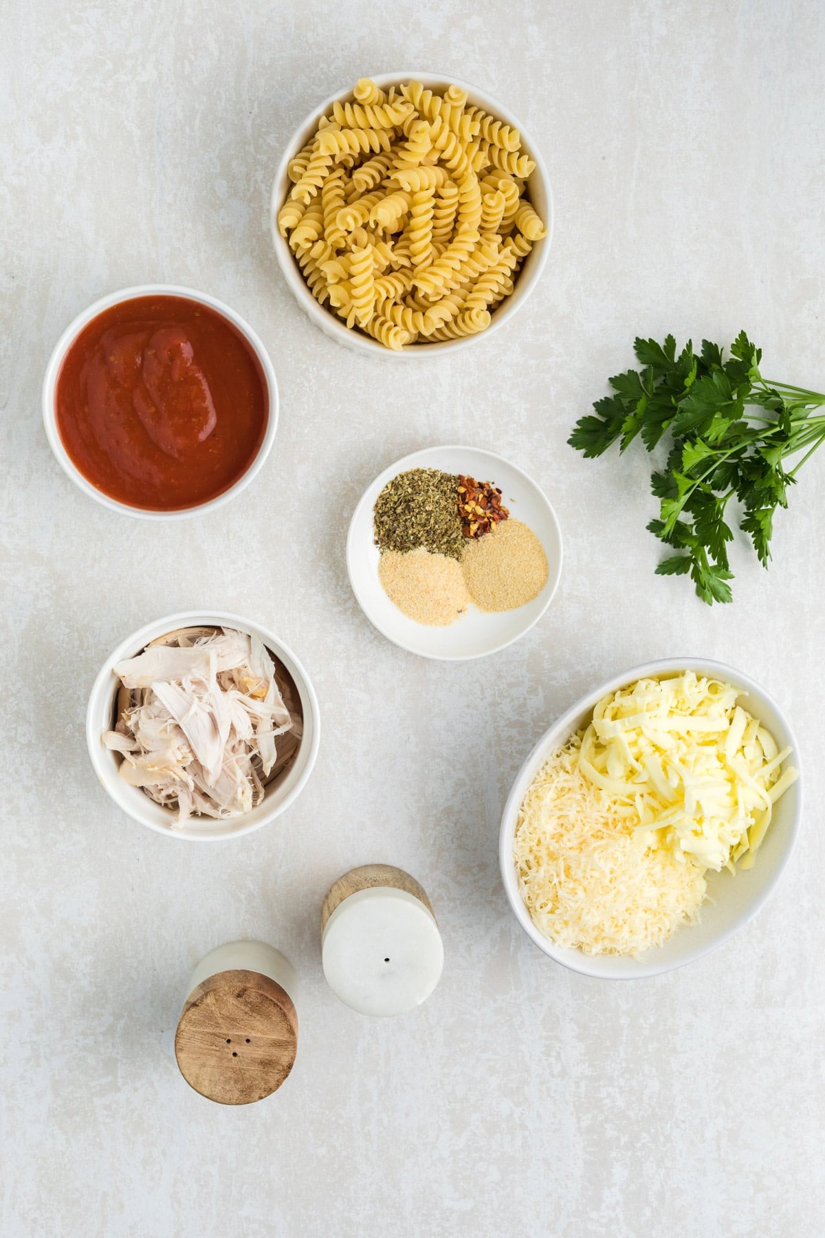 Flat lay of ingredients including rotini pasta, tomato sauce, shredded chicken, grated cheese, assorted spices, fresh parsley, and salt and pepper shakers on a light surface.