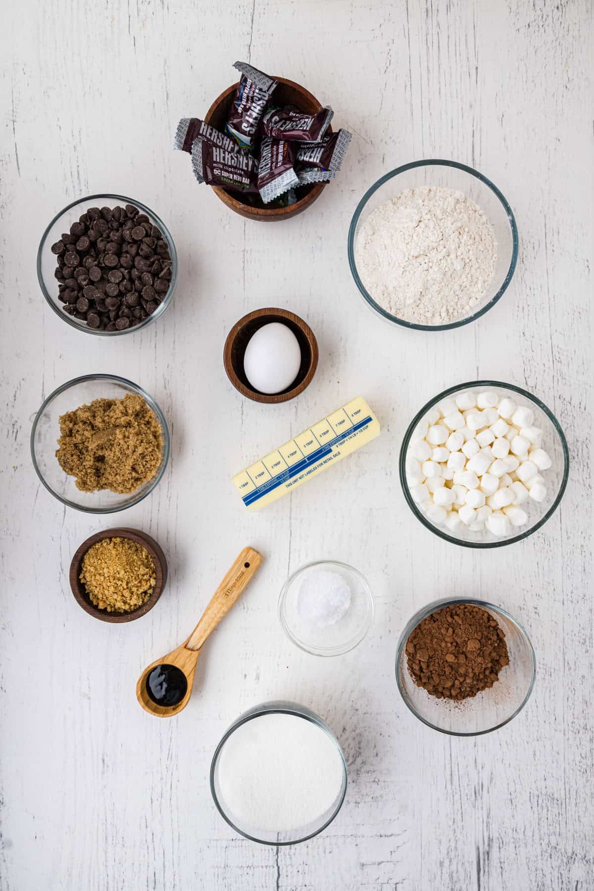 Various baking ingredients are arranged on a white surface, including chocolate chips, marshmallows, flour, sugars, cocoa powder, butter, an egg, and small chocolate bars.