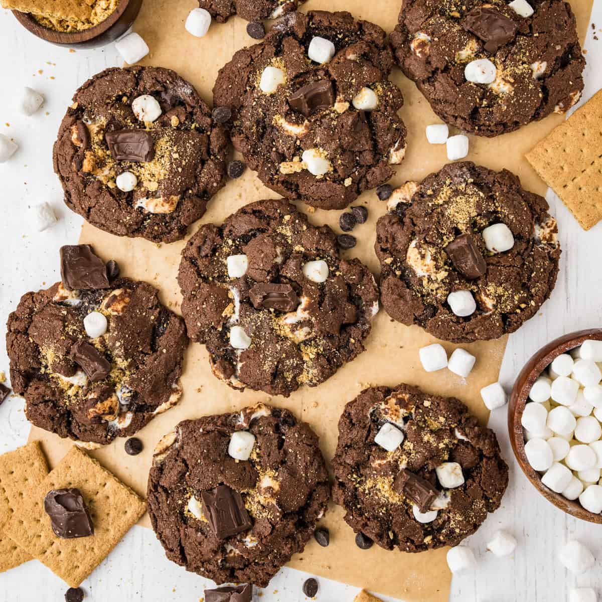 Nine chocolate s'mores cookies with marshmallows, chocolate pieces, and graham cracker crumbs are arranged on parchment paper, surrounded by mini marshmallows, chocolate chips, and graham crackers.