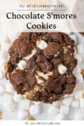 A chocolate smores cookie with marshmallows and chocolate chunks, surrounded by mini marshmallows. Text: Chocolate Smores Cookies and aweekendcook.com.