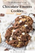 Two chocolate smores cookies with marshmallows and chocolate chunks rest on a cooling rack, surrounded by mini marshmallows and chocolate pieces.