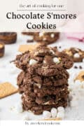 A stack of chocolate smores cookies with marshmallows and graham crackers on a white surface, surrounded by baking ingredients.