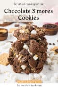 Two chocolate smores cookies with marshmallows and chocolate chips are stacked on a white surface, surrounded by graham crackers and mini marshmallows.