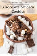 A bowl filled with Chocolate Smores Cookie pieces, surrounded by mini marshmallows, graham crackers, and chocolate pieces, with a recipe title and website name displayed.