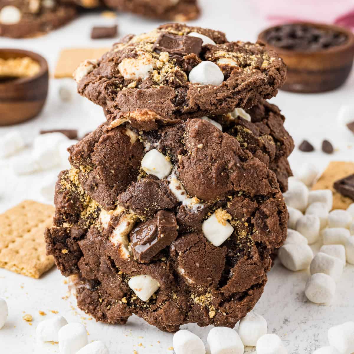 A stack of chocolate s'mores cookies with marshmallows, chocolate chunks, and graham cracker pieces surrounded by mini marshmallows and cracker pieces.