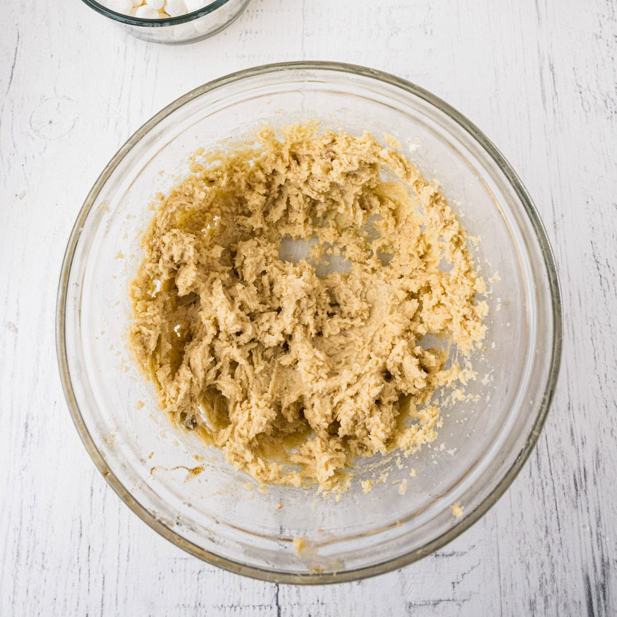 A glass bowl containing creamed cookie dough mixture sits on a white wooden surface.