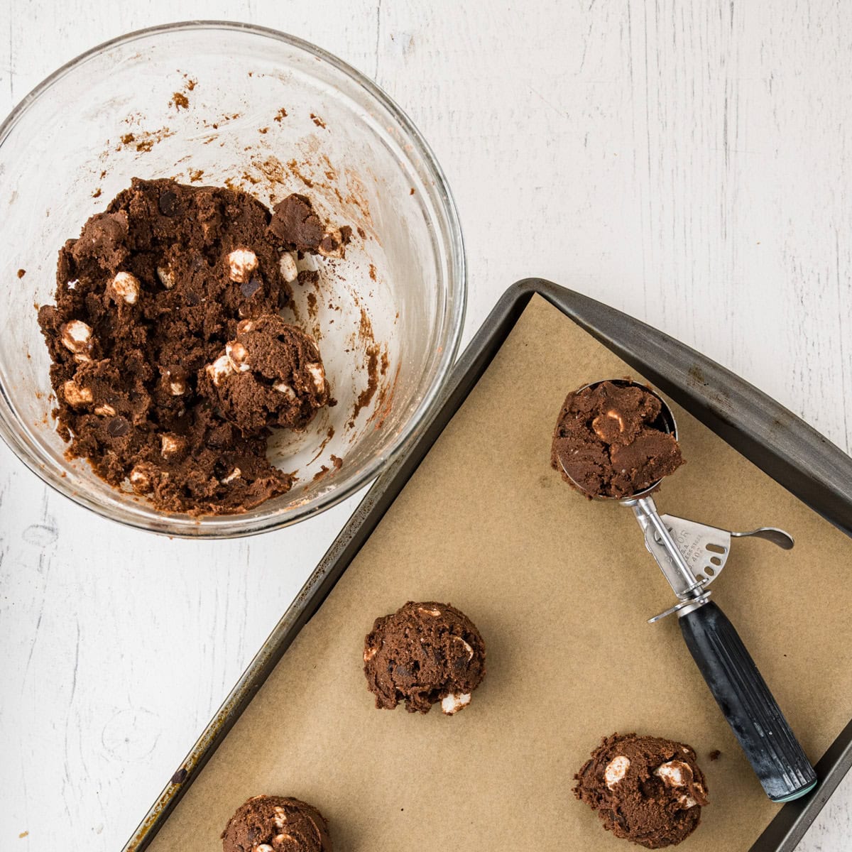 A glass bowl of chocolate s'mores cookie dough with marshmallows sits next to a baking tray with scoops of dough on parchment paper and a metal cookie scoop.