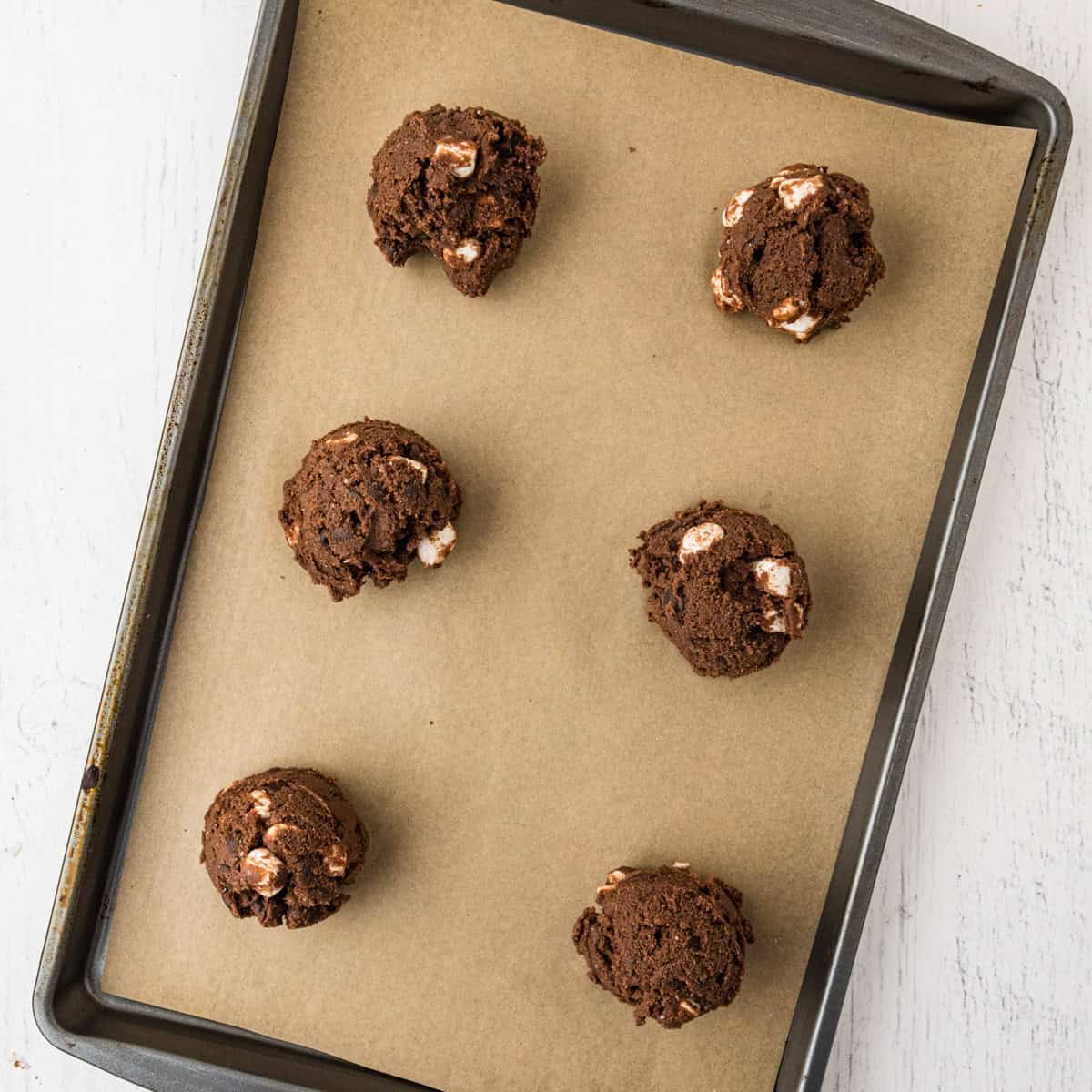 Six unbaked chocolate s'mores cookie dough balls with marshmallow pieces are arranged on a parchment-lined baking tray.