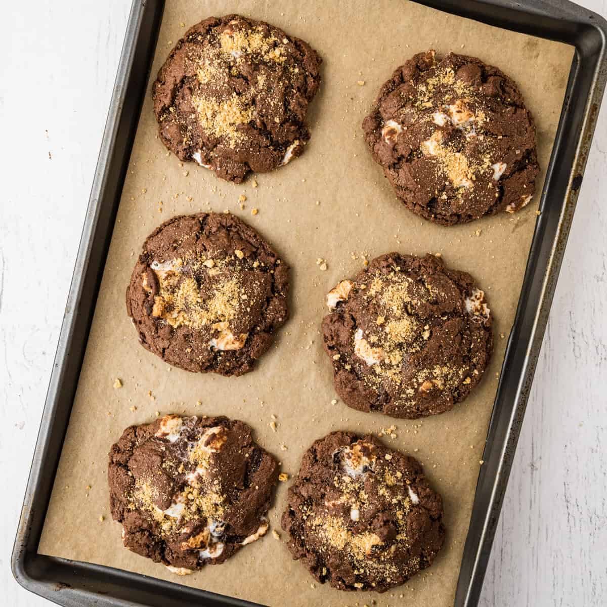 Six chocolate s'mores cookies with visible marshmallow pieces and a sprinkle of crumbs on top, arranged on a parchment-lined baking sheet.
