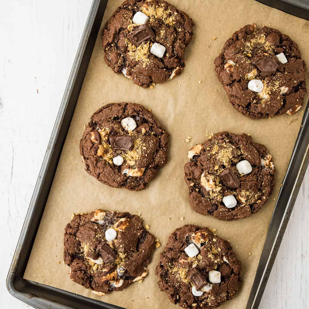 A baking sheet with six chocolate s'mores cookies topped with mini marshmallows, chocolate chunks, and graham cracker crumbs on parchment paper.