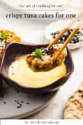 A tuna cake held with chopsticks is dipped into a creamy sauce, topped with black sesame seeds and a green garnish, with a small dish of more sesame seeds nearby.